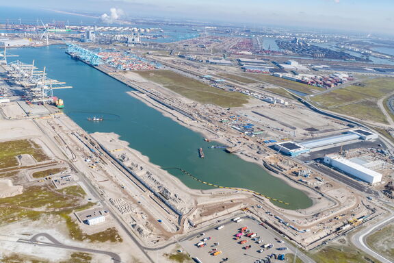Aerial view of a large port area on the Maasvlakte in Rotterdam, featuring container terminals, cranes, waterways, and industrial buildings. The image shows a wide channel surrounded by construction activity and infrastructure, with ships in the water and a vast industrial area in the background.