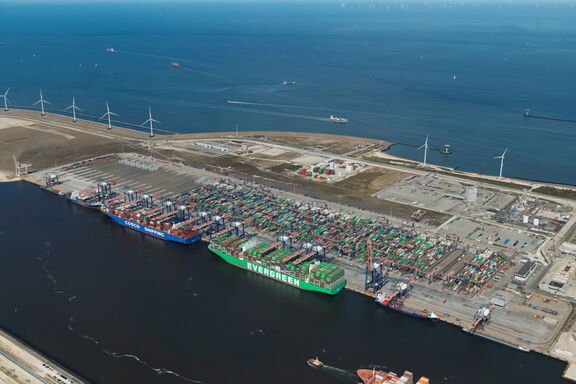 Aerial view of the Euromax container terminal on the Maasvlakte in Rotterdam, with large container ships from Evergreen and COSCO moored at the quay. The site is stacked with colorful containers and equipped with tall container cranes. Wind turbines and the North Sea are visible in the background, along with a few ships at sea.