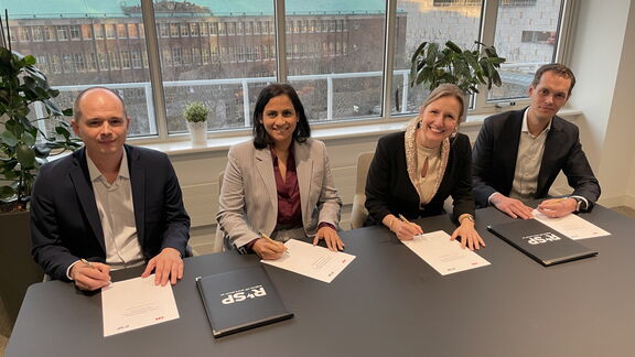 Four people are sitting at a table in a modern meeting room signing official documents. On the table are folders and papers bearing the “RSP” logo. In the background is a large window overlooking urban buildings, including one with the inscription “ABN AMRO.” There are also a few plants on the windowsill.