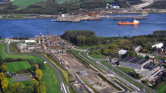 An aerial photograph showing a large construction site adjacent to a river. Several cranes and heavy machinery are visible on the site, along with organized work areas. Across the river, there are industrial structures and a docked orange cargo ship. Surrounding the area are green fields, roads, and buildings, indicating a mix of industrial and rural landscape.