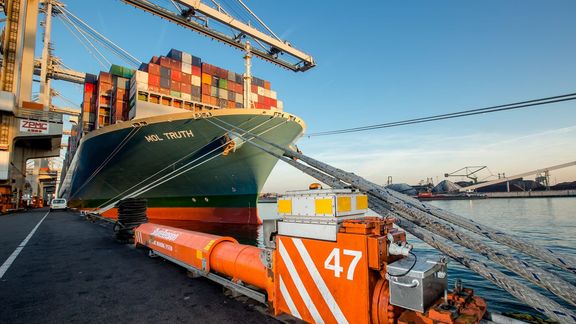 Containerschip ligt afgemeerd aan een terminal in de haven, waar kranen containers laden en lossen.