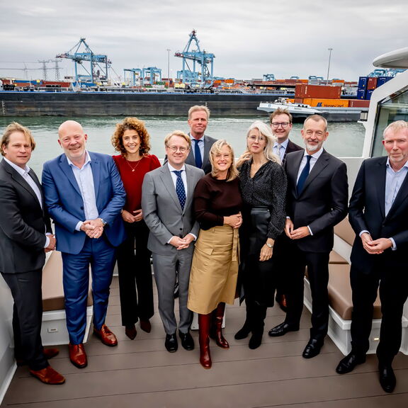 A group of people are standing together on the deck of a boat in a harbour setting. Containers, cranes and ships are visible in the background, indicating an industrial harbour location. The individuals are neatly dressed in business attire, including sui